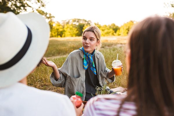 Pretty girl holding juice in hand thoughtfully discuss something with friends spending time on picnic in park